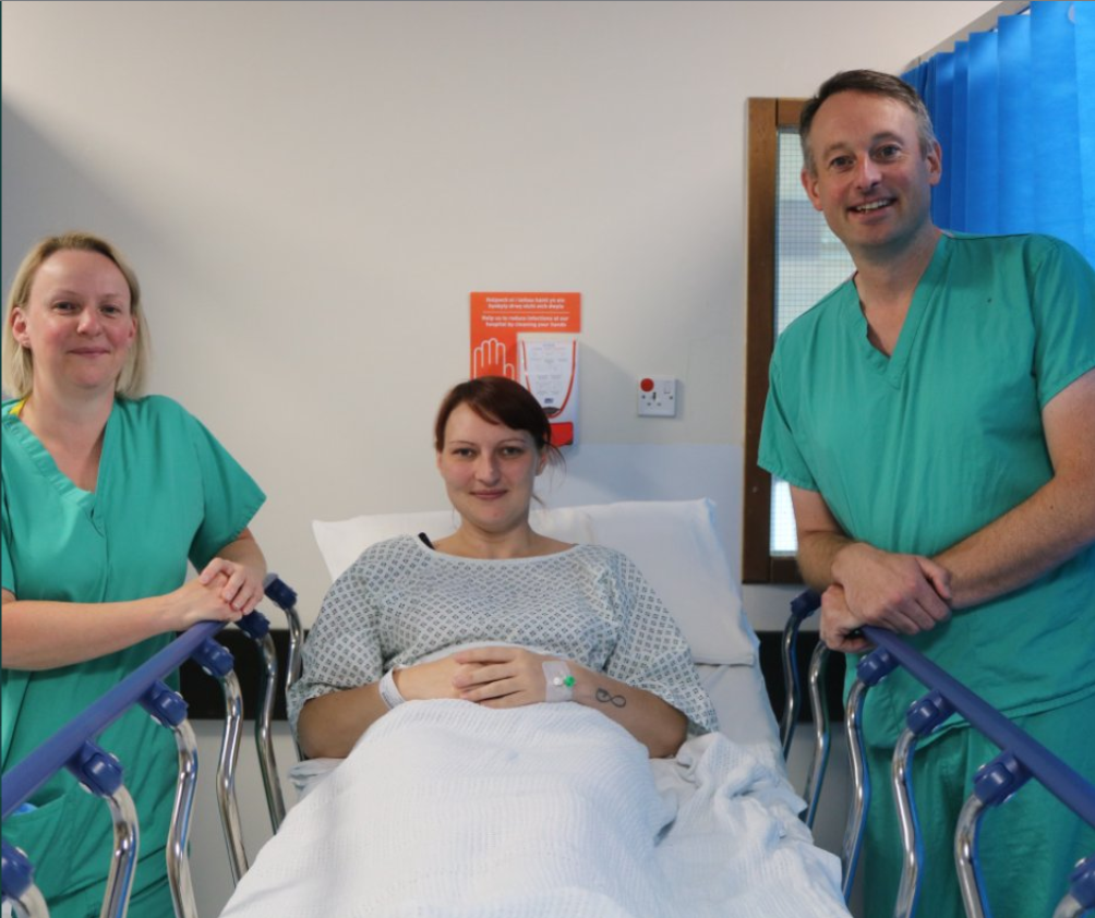 Patient of first robotic assisted hysterectomy in Wales, lying in hospital bed with doctors either side of her.