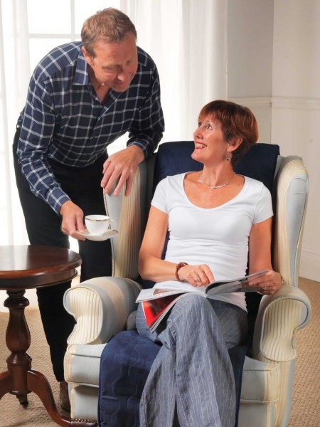 A blue seat warmer is on a cream armchair. A woman is seated on the seat warmer. There is a man to her left passing her a teacup. They are smiling at each other.