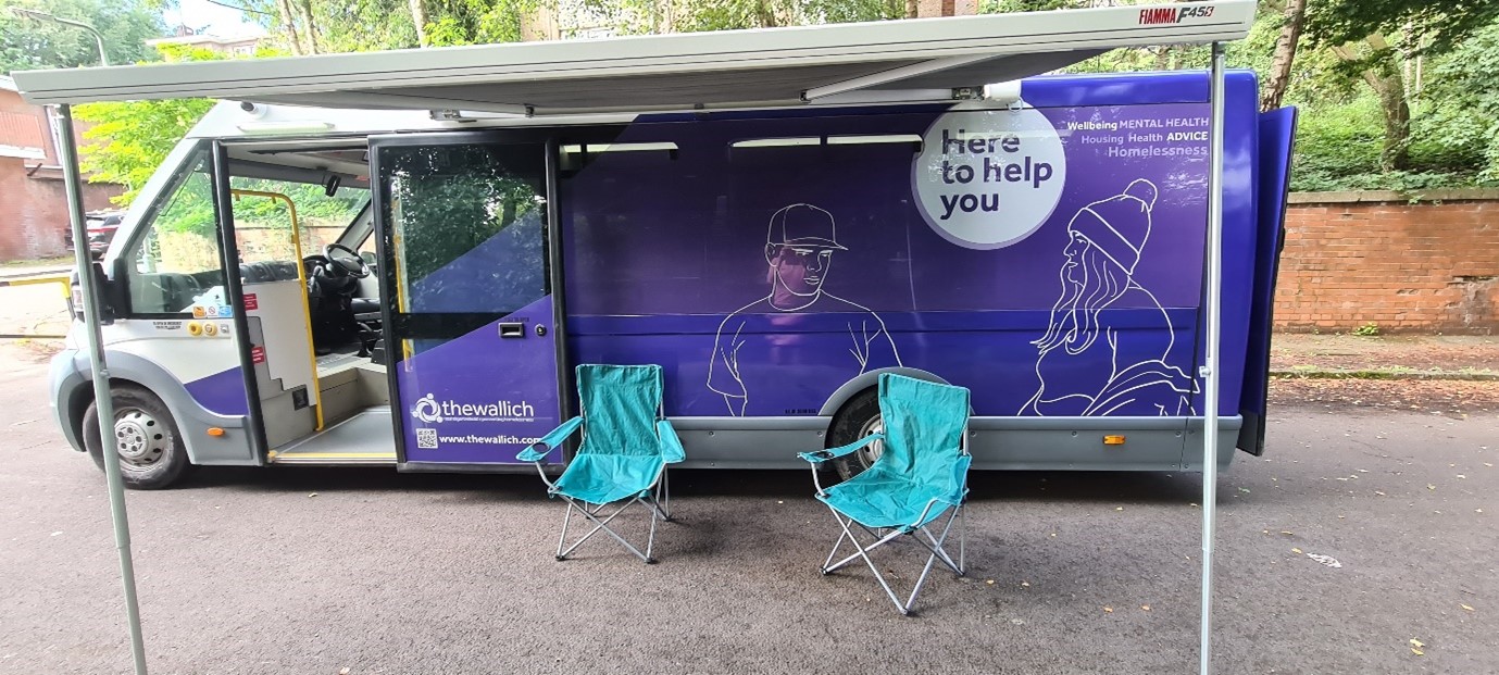 The Wallich bus is parked and has a canopy extending from the vehicle. Two chairs are underneath the canopy.