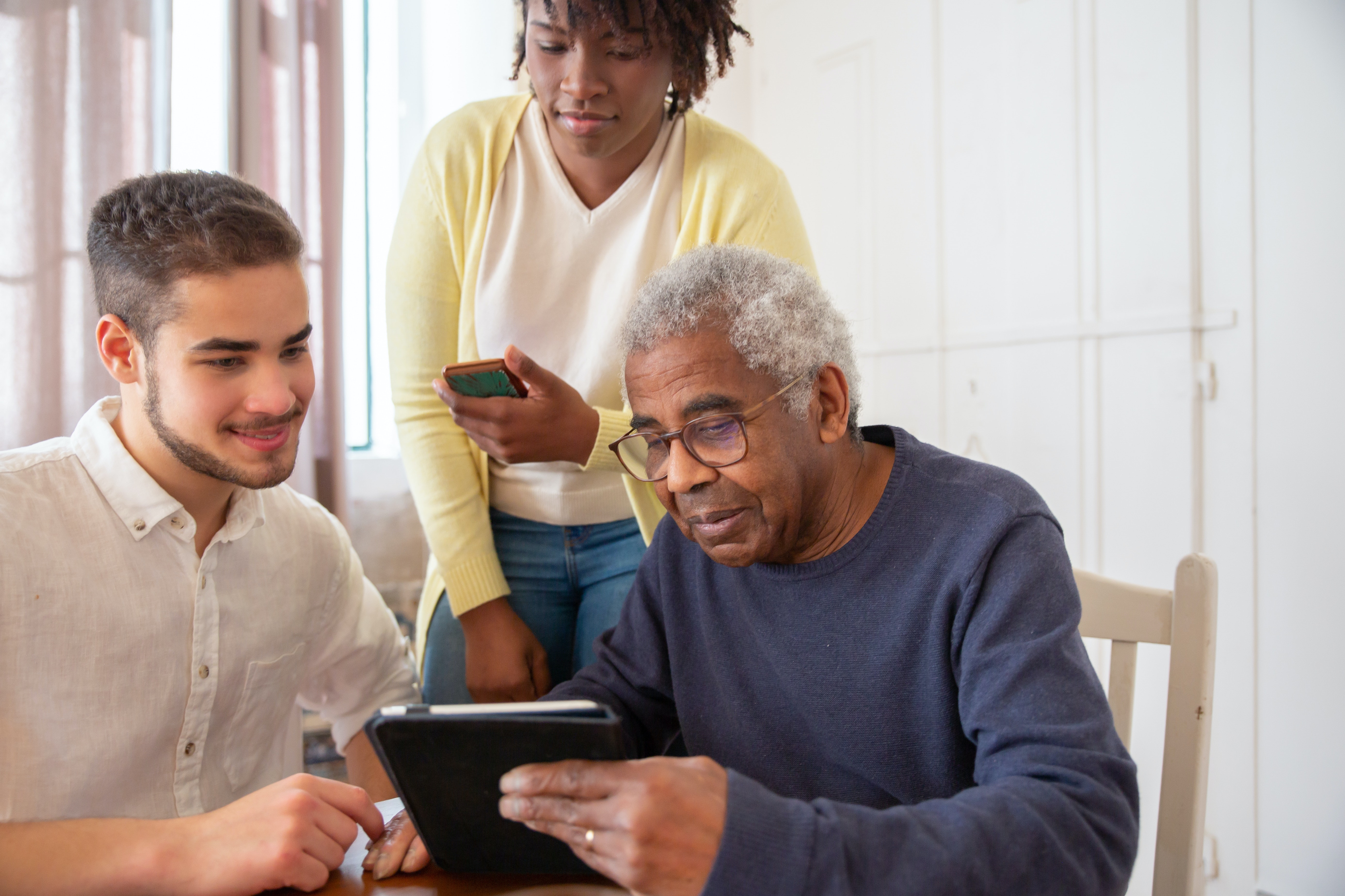 an elderly man holding a smart tablet with two people watching beside him