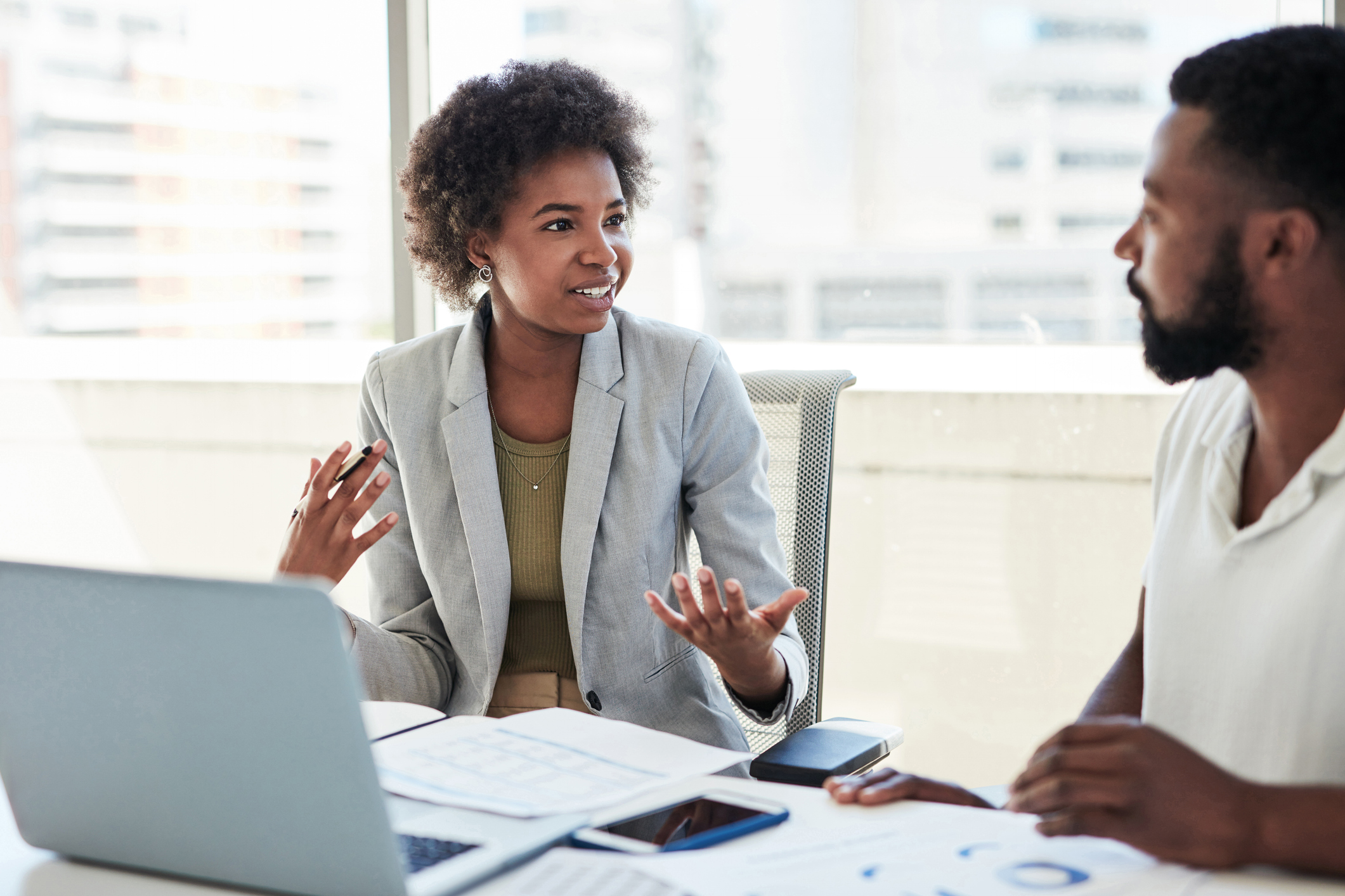A woman explaining something to a man. They are smartly dressed and sat in front of laptops and paperwork.