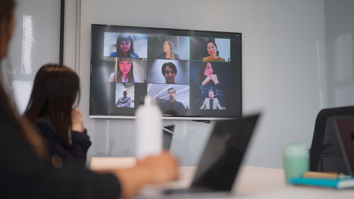 People sitting at table with laptops in front of them. They are all looking at a big screen with a group of people on it.