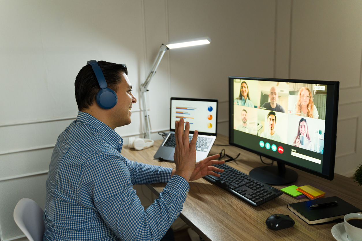 Man sitting at desk on video call. He's waving at his colleagues on screen.