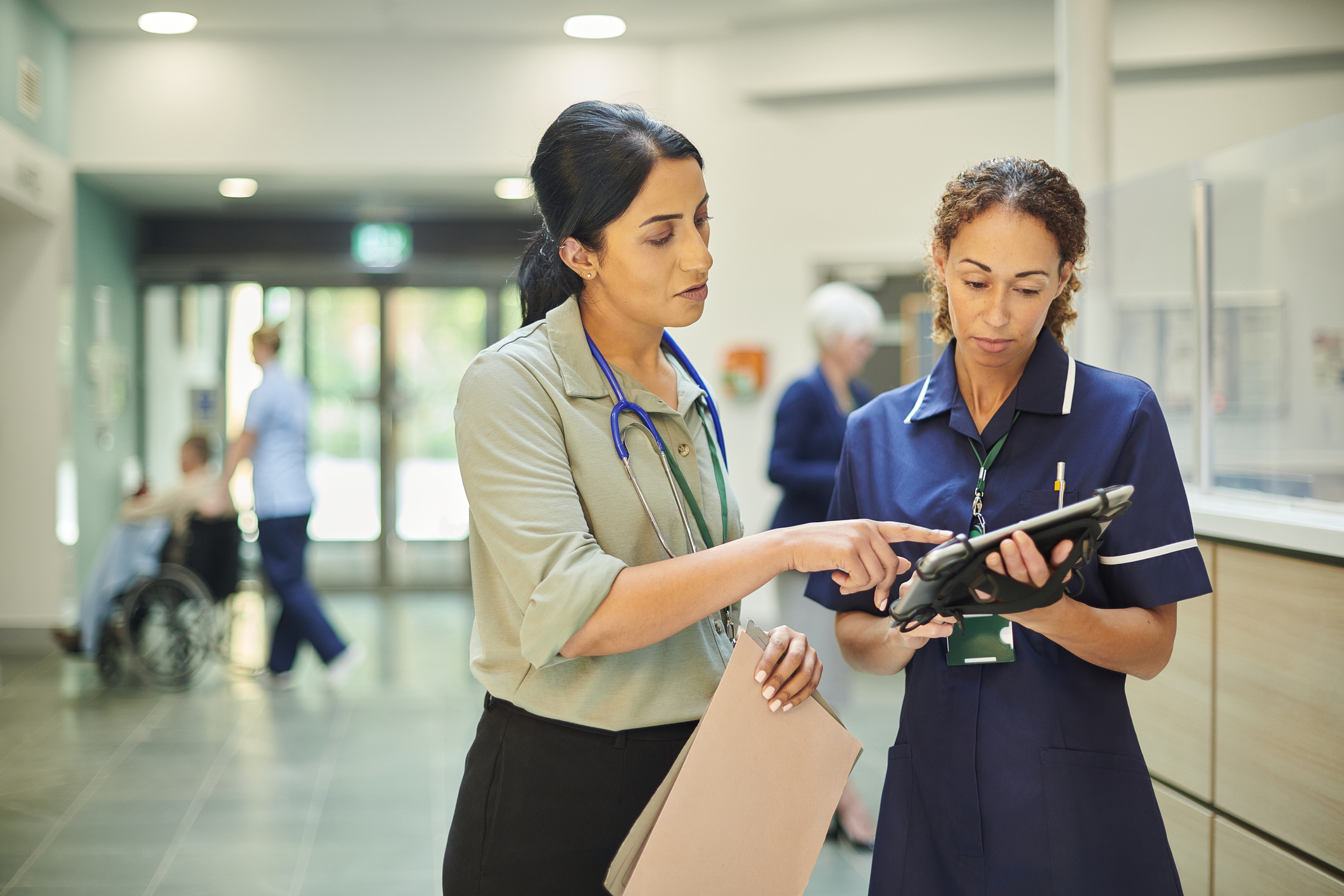 Two women are in a hospital corridor. One has an iPad and the other is pointing at it. One is dressed with a stethoscope around her neck whilst the other wears a nurse uniform.