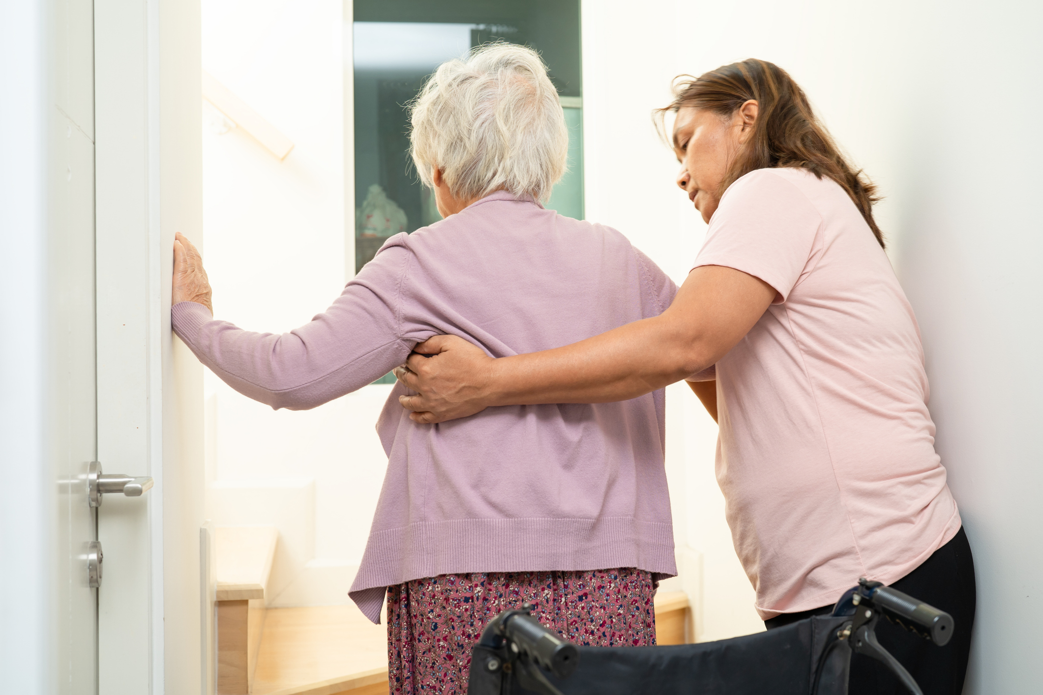 Elderly woman standing up out of a wheelchair. A carer is helping her stand. Both are facing away from the camera.