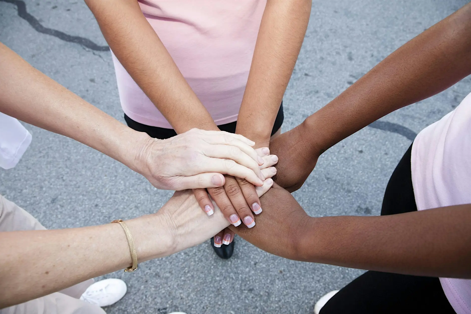 Three women stand in a circle. They are reaching out both hands and placing them on top of each others hands.