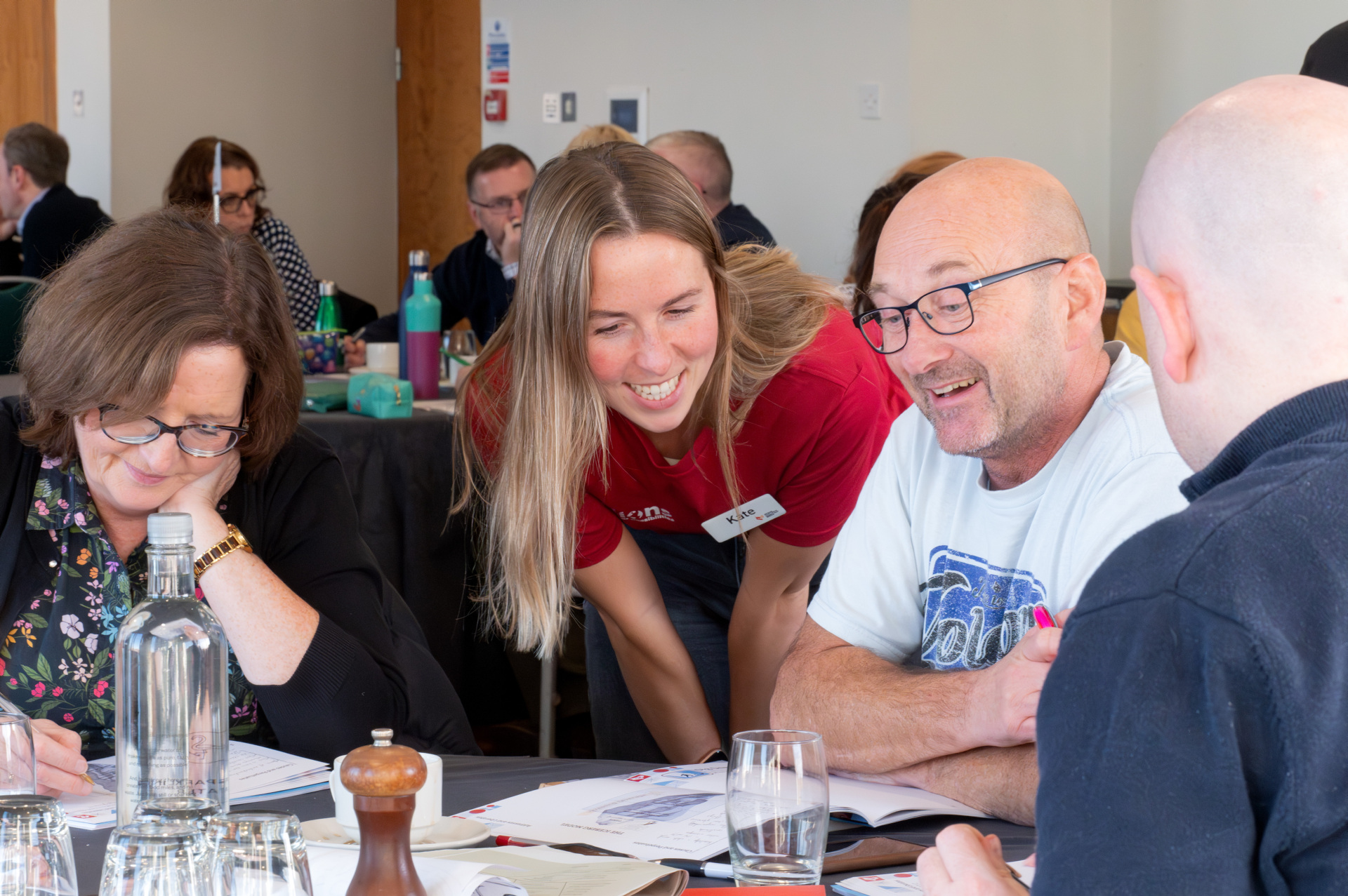 A group of people are sitting at a table. A woman is bent over someone's shoulder contributing to the discussion. 