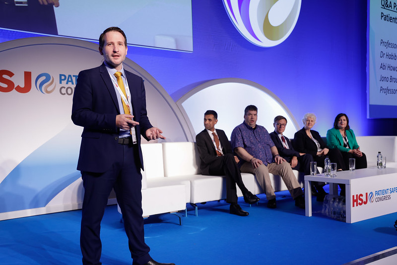 Man on stage talking to audience. Behind him there are five people sat on a sofa. They're a panel who are about to present.